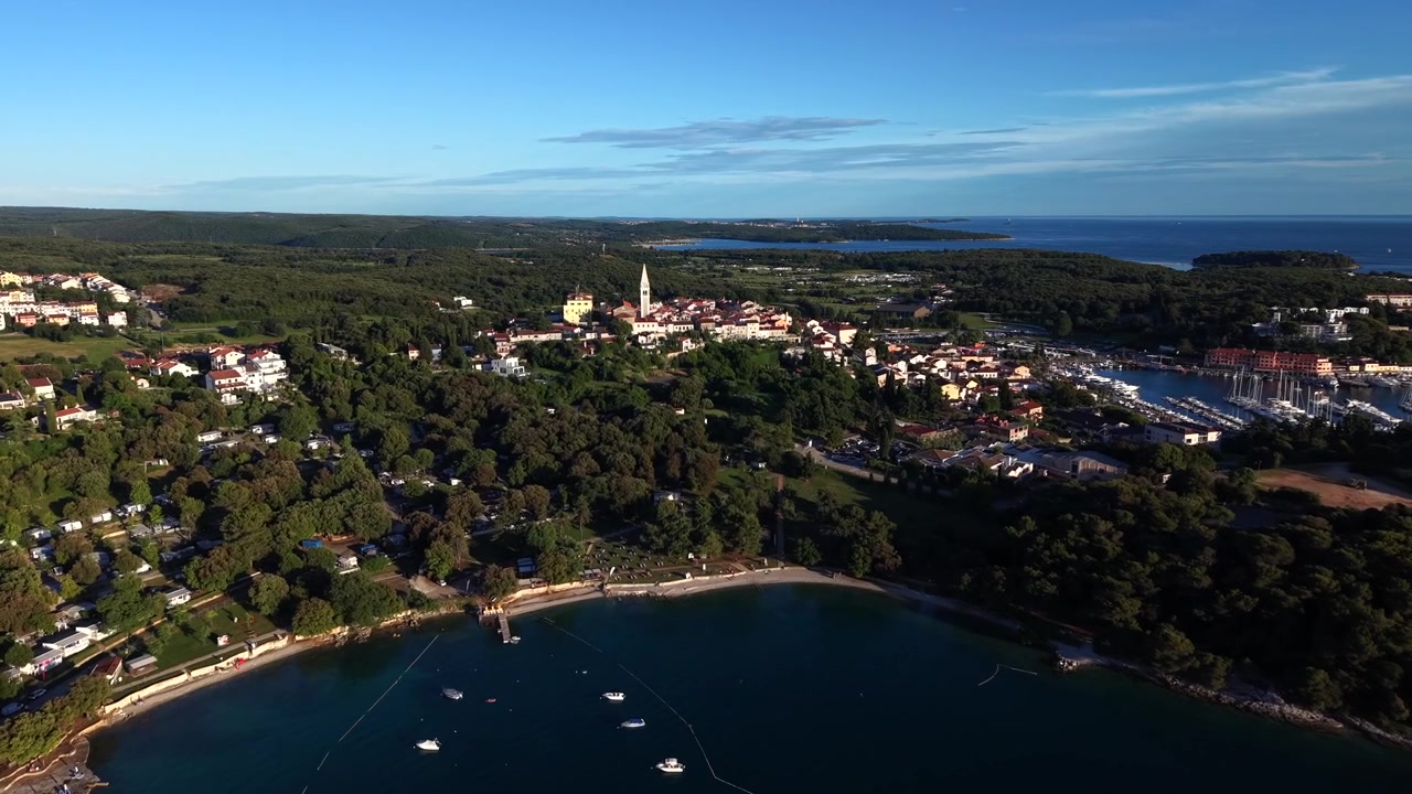 Storica città collinare di Vrsar con campanile della chiesa si affaccia su una baia tranquilla punteggiata da piccole imbarcazioni, un porto turistico e una costa ricoperta di pini alla luce del primo mattino. Croazia. Ripresa aerea con drone in avvicinamento.