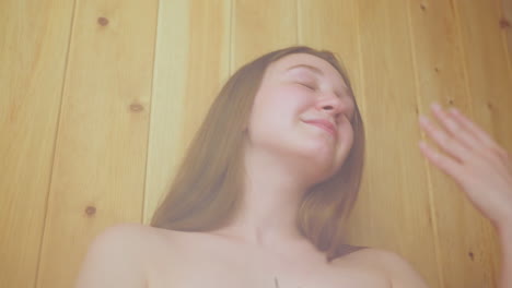 elegant woman wrapped in towel relaxes inside wooden sauna, gently fanning herself with hand while enjoying soothing heat and steamy air, soft smile on face, surrounded by warm light