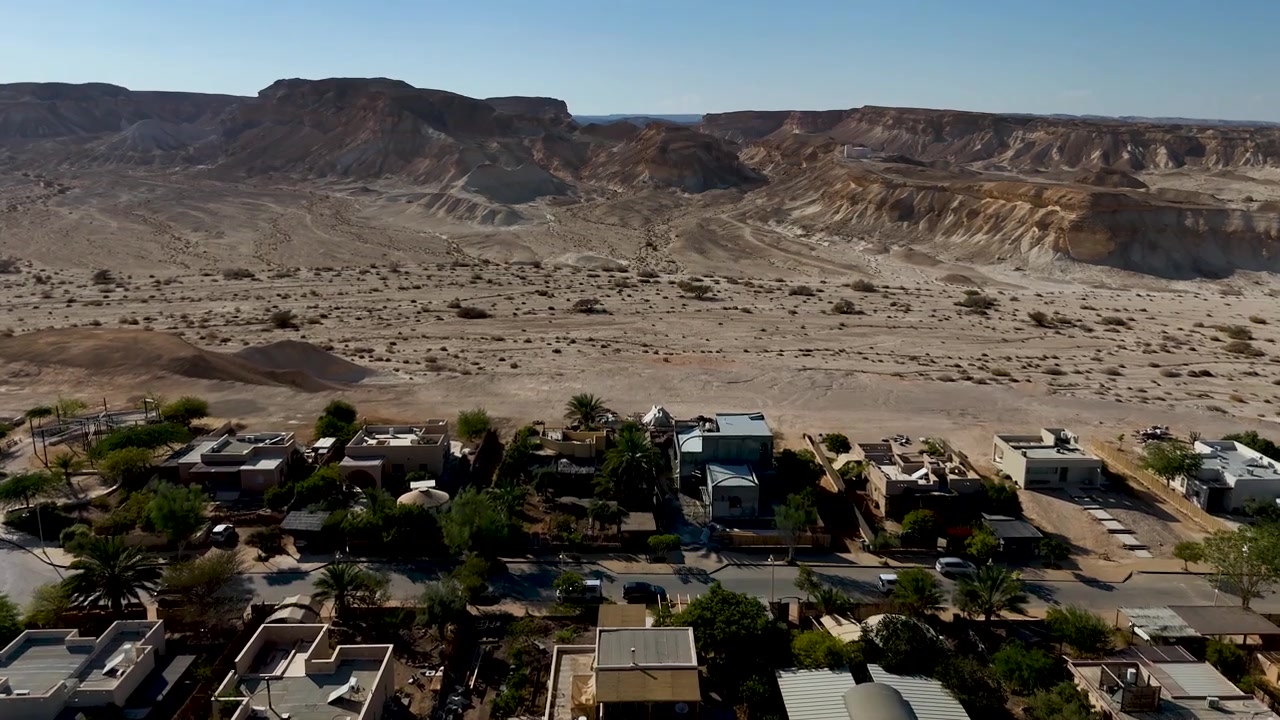 4K 60fps drone footage flying west along the edge of Zukim village, Israel, gradually tilting downward toward a secluded villa surrounded by the rugged Arava Desert landscape