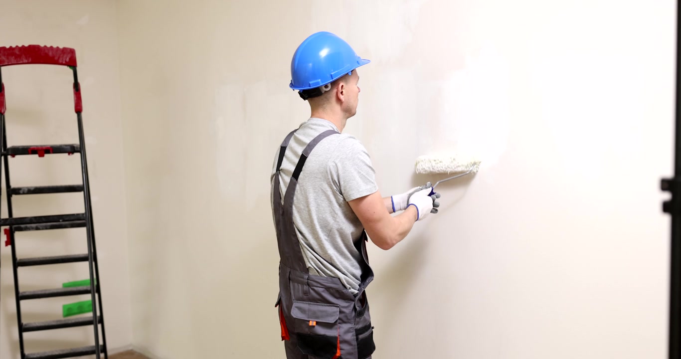Worker in helmet paints wall with roller brush in light room