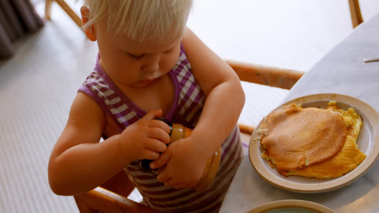 Toddler grabbing jar at table showing code scroll, twisting lid, drizzling spread onto pancakes