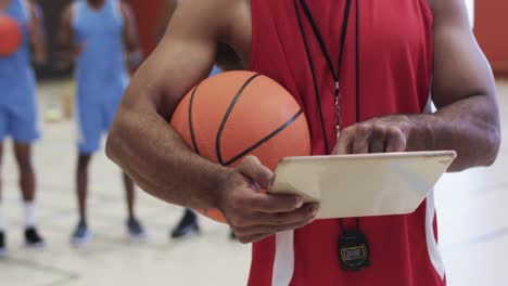 midsection of biracial male basketball coach using tablet at indoor court, in slow motion