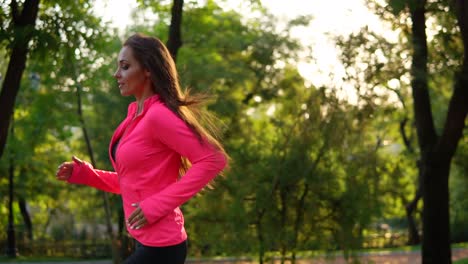 Young-woman-in-bright-pink-jacket-running-in-the-sunny-city-park-exercising-outdoors.-Steadicam-stabilized-shot,-Slow-Motion