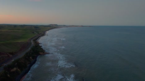 Vista-Aérea-Por-Drones-Del-Tráfico-Que-Viaja-A-Lo-Largo-De-La-Carretera-Costera-Con-Vistas-Al-Océano-Y-Un-Hermoso-Resplandor-Naranja-En-El-Cielo-En-La-Península-De-Katiki,-Otago-Del-Norte,-Nueva-Zelanda-Aotearoa