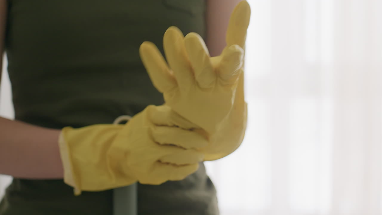 Close up of cleaner in yellow gloves adjusting fit by stretching fingers while preparing for cleaning duty indoors with blurred bright background