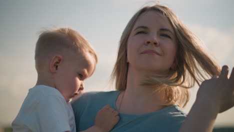 close-up of a woman in a blue dress carrying her young son, who is wearing a white top. the boy looks sad while the woman gently moves her hair off her face