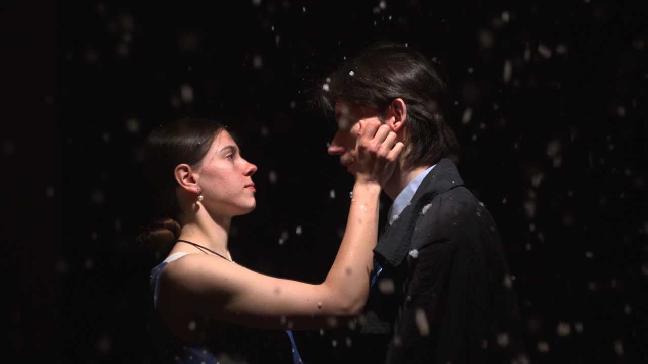 A man on the right faces a woman on the left in a dark studio, gently touching his hat while snowlike particles drift through dramatic low light