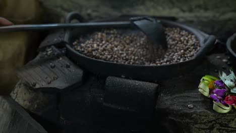 burning log, charcoal and flame heating up the iron pan for traditional bali roasting of luwak coffee with wooden spoon in presence of rising steam and hindu flower offering