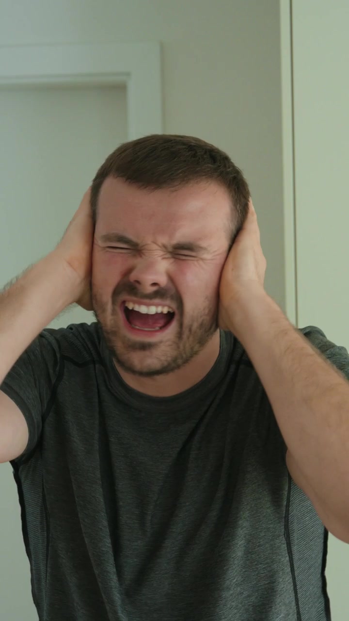 Close-up shot of a man covering his ears with an annoyed expression, showing noise sensitivity, stress, refusal to listen, frustration, overload, conflict, and emotional reaction.