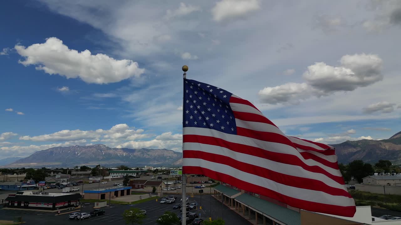 Bandiera americana USA che sventola al vento in una bella giornata di sole estiva con nuvole e cieli azzurri che si affaccia su una piccola città americana USA e montagne mentre il drone vola e ruota intorno all'asta della bandiera - in 4K a 60fps