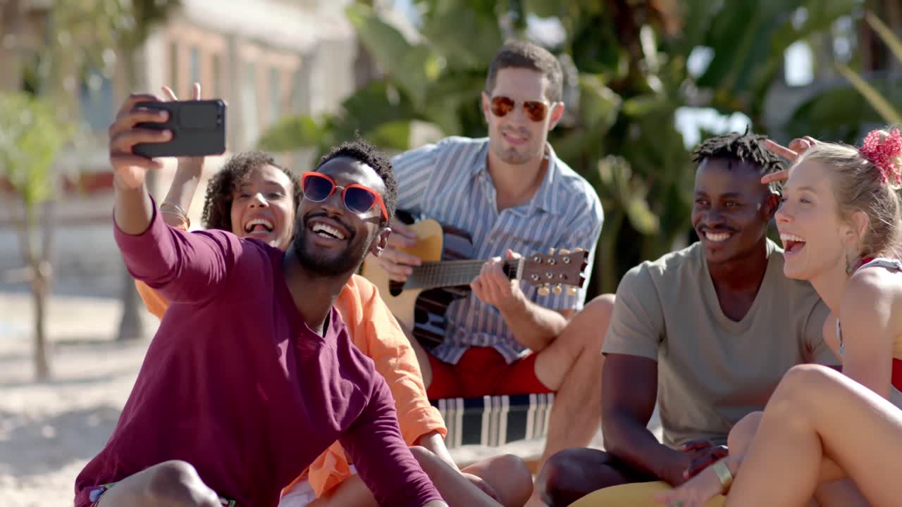 un grupo feliz y diverso de amigos tomando selfies y tocando la guitarra en la playa con palmeras