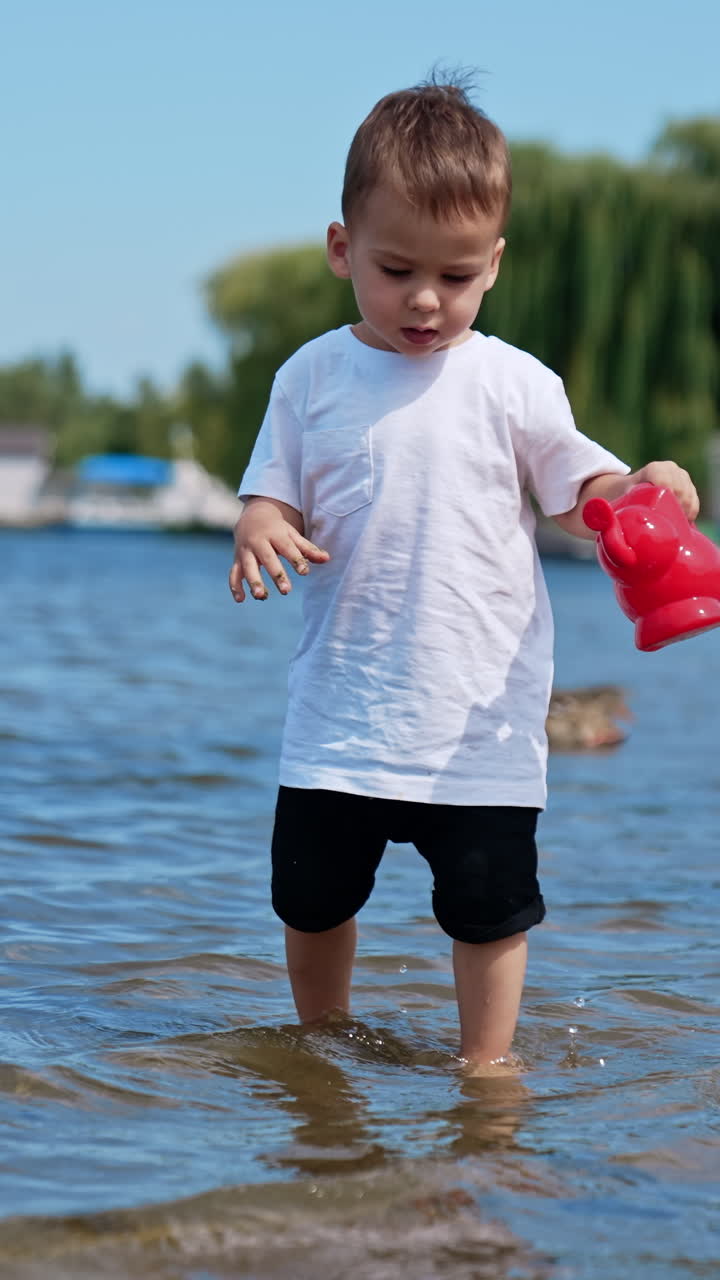 Cute baby boy plays with a watering can. Toddler tries to fill the can. Child playing in the beach. Vertical video