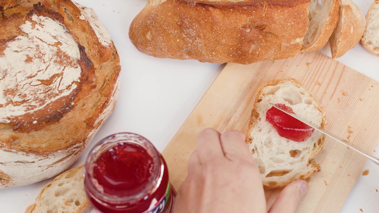 Overhead view of hand scooping strawberry jam from jar onto crusty sourdough bread slice.