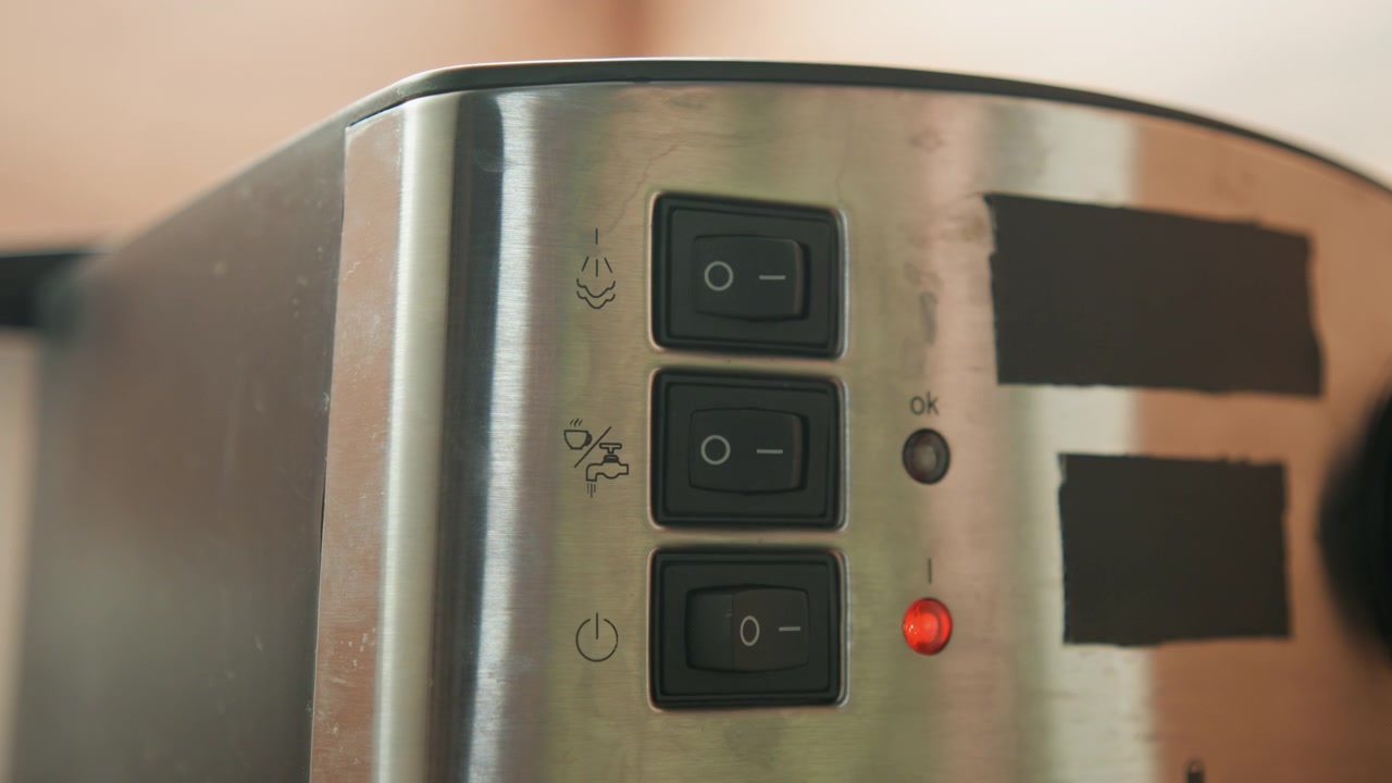 Horizontal close-up shows stainless steel toaster control panel with three black rectangular buttons, fingertip pressing one button and small red indicator light glowing on metal surface