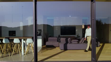 a drone captures the progress of a man from the living room to the kitchen through the large glass windows of the house