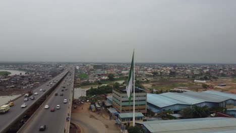 Coches-Moviéndose-Rápidamente-En-El-Puente-De-Una-Carretera-Suburbana-Con-Una-Bandera-Nigeriana-Frente-A-Una-Industria.