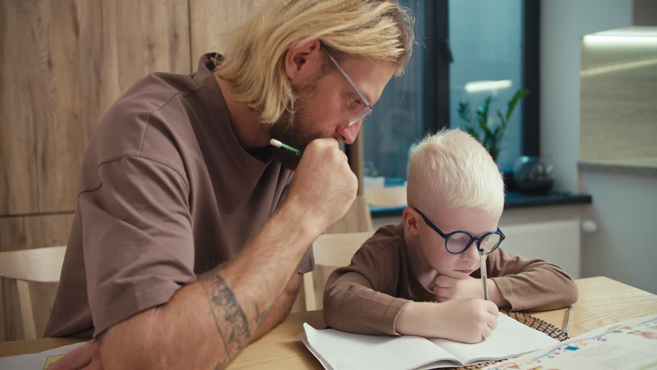 A little albino boy with white hair in blue glasses holds a pen in his hands and writes something in his notebook. His father, a blond man, helps him do his homework