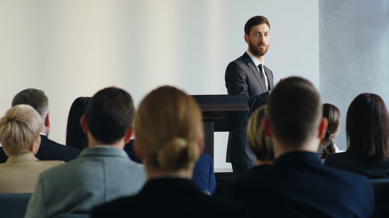 Caucasian businessman speaker on a podium wearing formal clothes and talking in a conference room in front to many people
