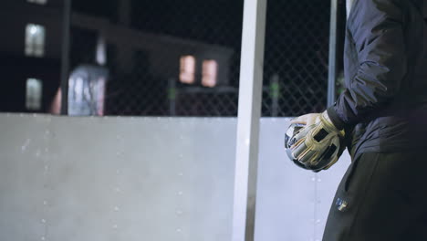 primer plano de un atleta con guantes pateando una pelota de fútbol bajo luces nocturnas durante la sesión de práctica, el fondo incluye edificios residenciales iluminados y un entorno deportivo urbano