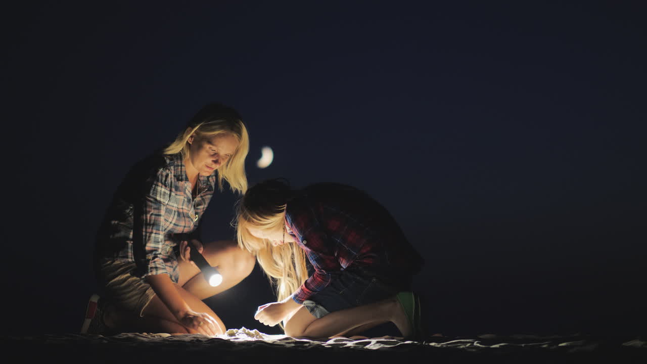 mamá e hija están jugando juntas en la arena por la noche brillan con una linterna buscando