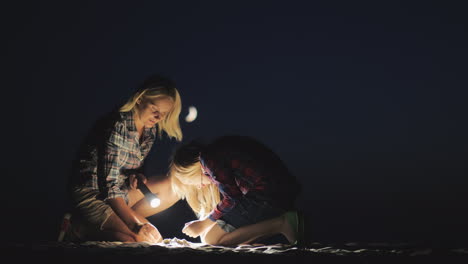 mom and daughter are playing together in the sand at night they shine with a flashlight looking for