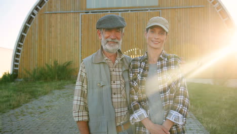 portrait of gray-haired caucasian old man with young woman standing outdoor at stable and smiling at camera on a sunny day