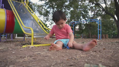 dolly en cámara lenta dentro y fuera del clip de un niño del este de asia que se divierte y sonríe jugando con juguetes sentados en el suelo de un parque infantil al aire libre