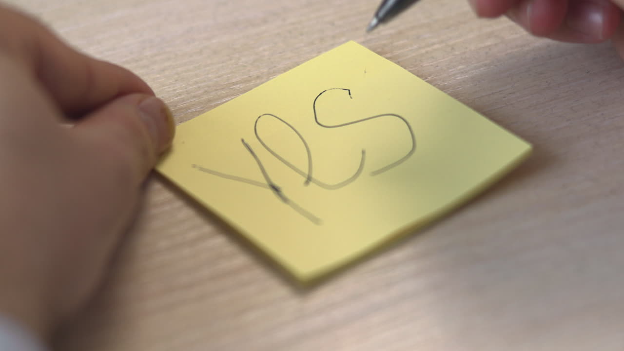Nærbilde av hender som skriver et tydelig 'Ja' på en gul post-it-lapp, bestemte pennestrøk, tekstur av trebord. Media.