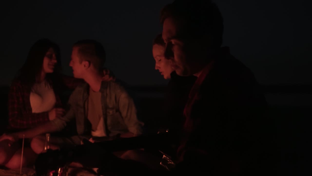 젊은이들이 불 근처에 앉아서 함께 노래를 부르고, 기타를 연주하고 있습니다. 피크닉, 불, 밤