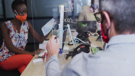 Rear-view-of-man-wearing-face-mask-writing-on-glass-board-while-sitting-on-his-desk-at-modern-office