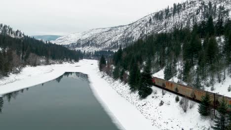 close-up of freight train traveling through a snowy landscape in british columbia: aerial shot follows journey along the partially frozen north thompson river and yellowhead highway 5 near kamloops