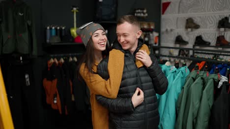 young man trying at winter jacket in clothing store with his girlfriend