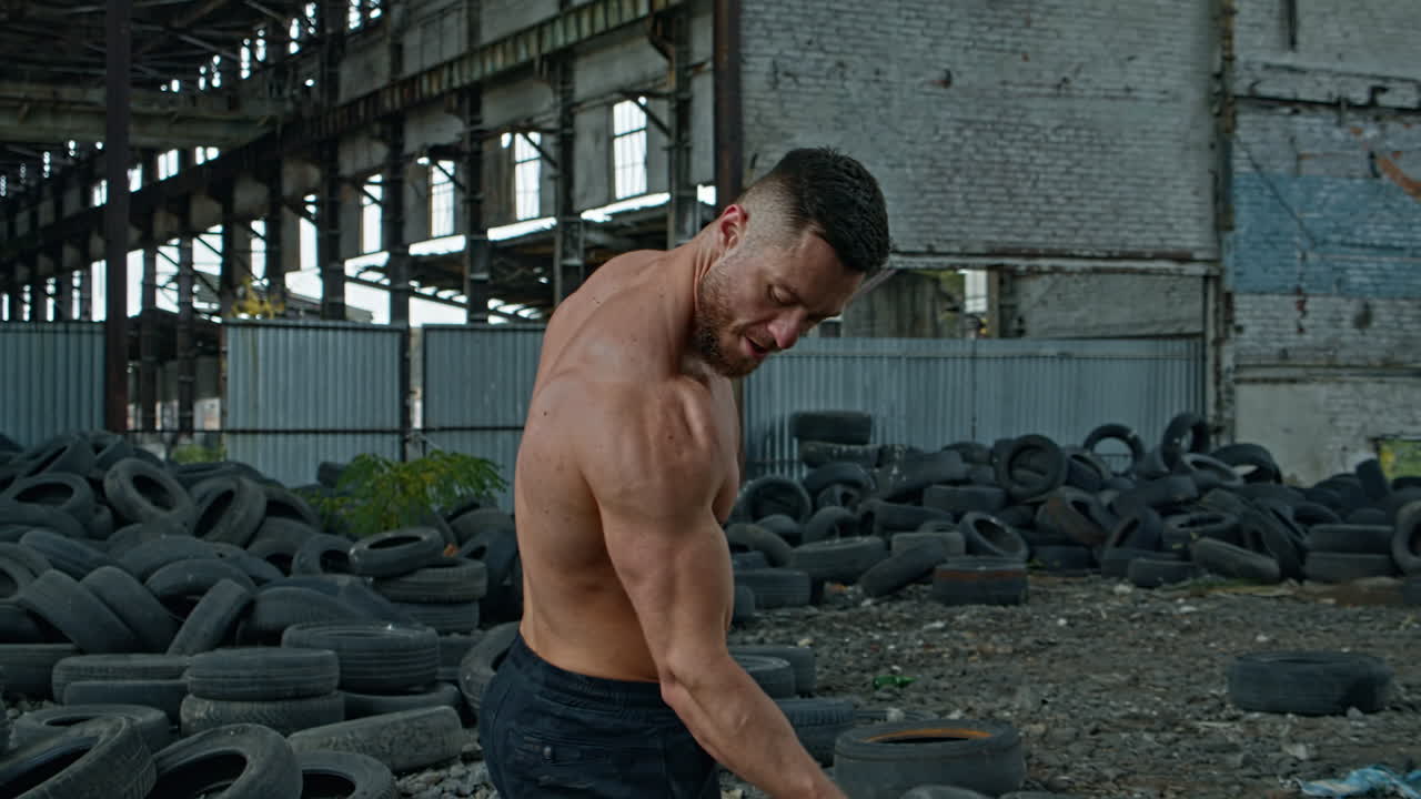 Male athlete with tires in abandoned place. Strong young man training his arm muscles with car tyre during an intense training session on ruined building background.