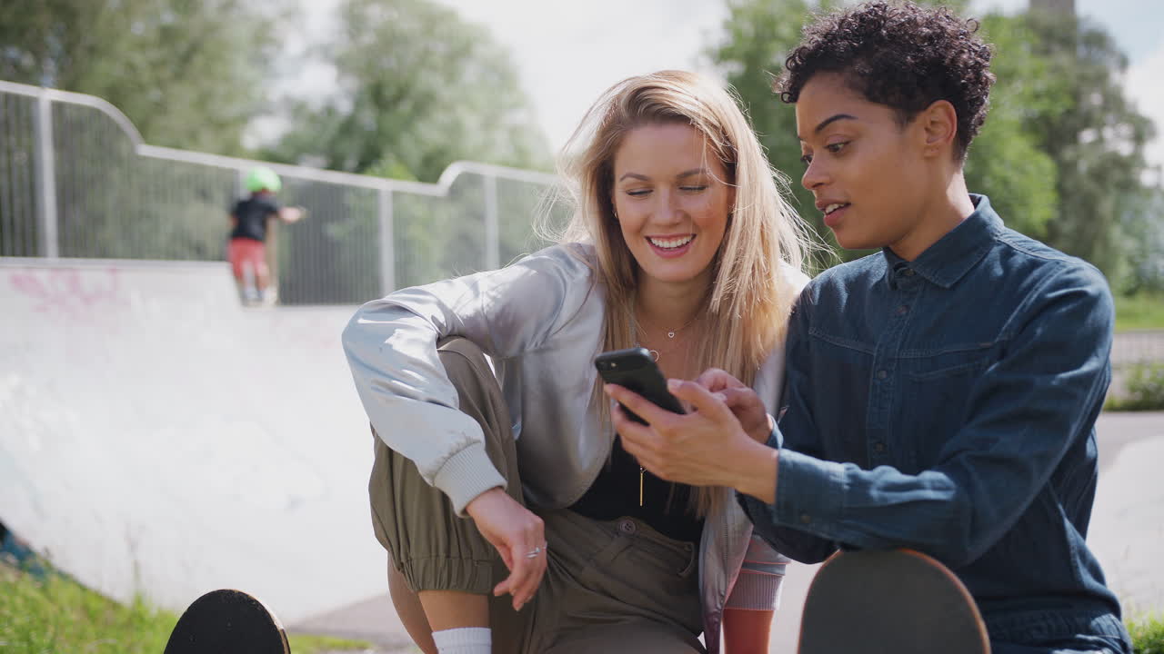 Two Female Friends Looking At Mobile Phone In Urban Skate Park And Laughing