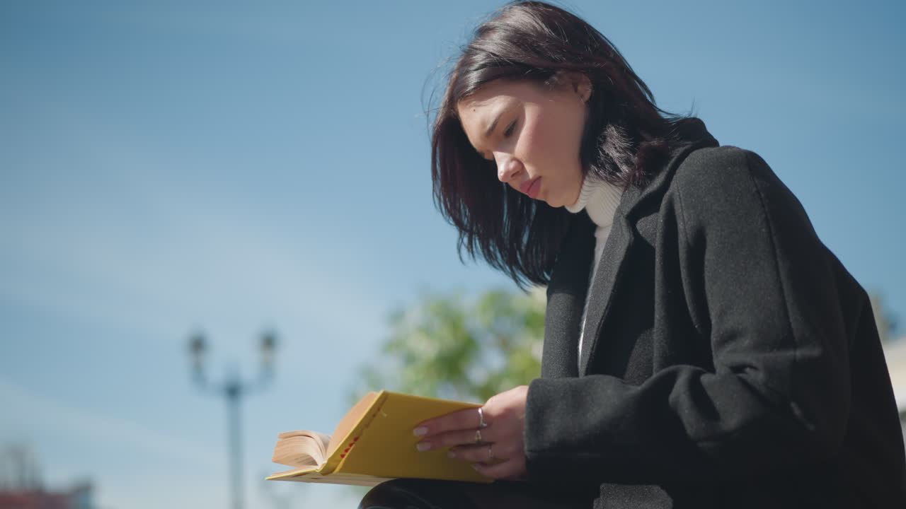 Close-up side view of student flipping pages of book outdoors. Wind gently fluttering hair while adjusting laptop, behind her is a red brick building in the background on a clear day