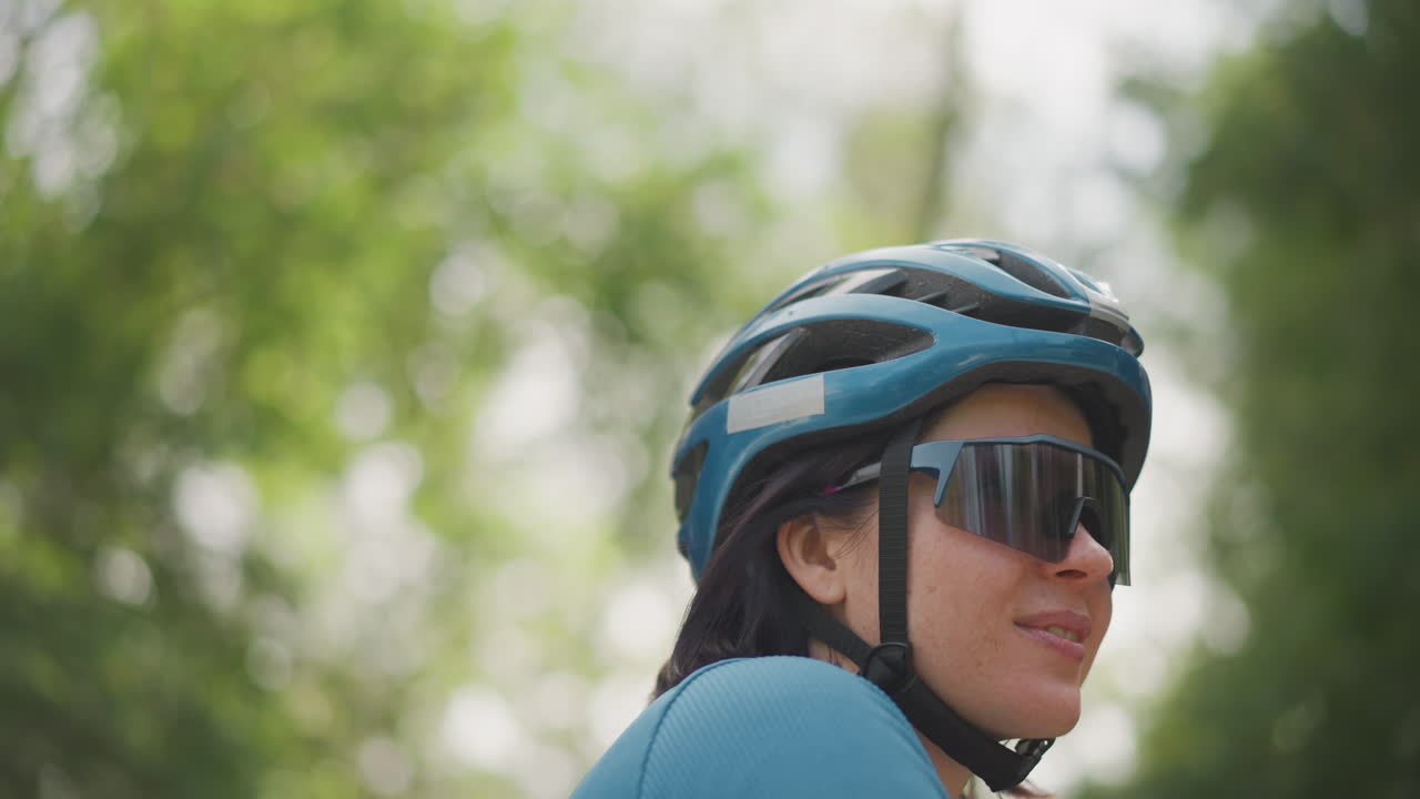 woman cyclist in blue helmet relaxed smile reflecting sunlight through trees, oversized sunglasses, side profile portrait, blue jersey on park trail, solo ride blending training and leisure, urban