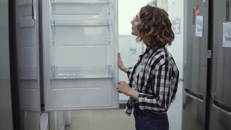 cheerful young girl selecting domestic refrigerator in supermarket. curly woman in plaid shirt deciding to buy a new one for home