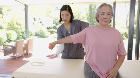 helping senior asian woman with arm exercise, physical therapist using small ball at home
