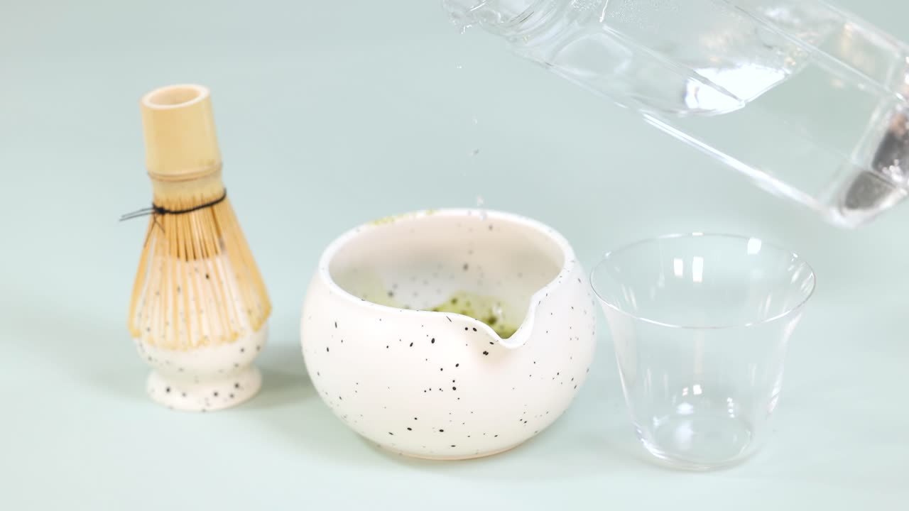 Clear water pours into a ceramic matcha bowl beside a bamboo whisk under soft lighting