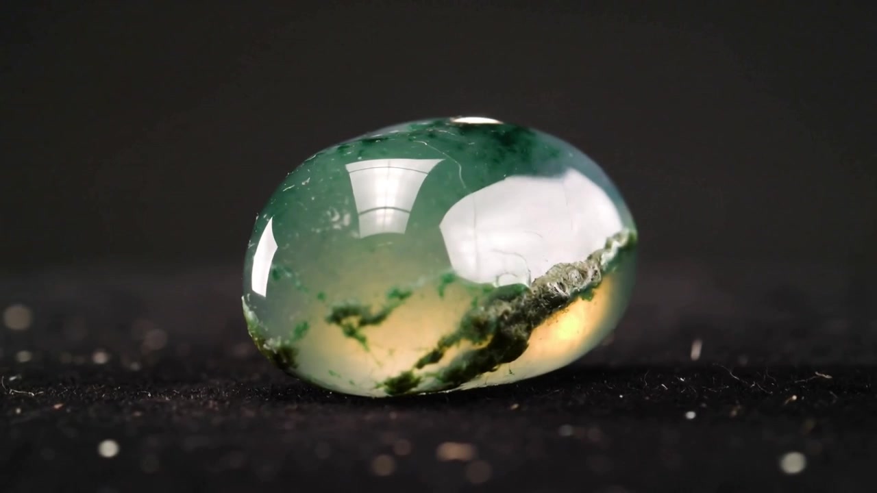 Cabochon verde in sottile rotazione che rivela dendriti muschiosi su un tappetino da studio scuro sotto una luce rettangolare