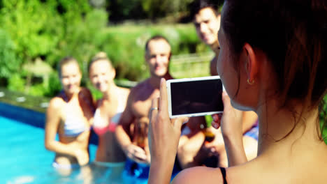 Mujer-Sonriente-Haciendo-Clic-En-Fotos-De-Amigos-Desde-Un-Teléfono-Móvil-Cerca-De-La-Piscina