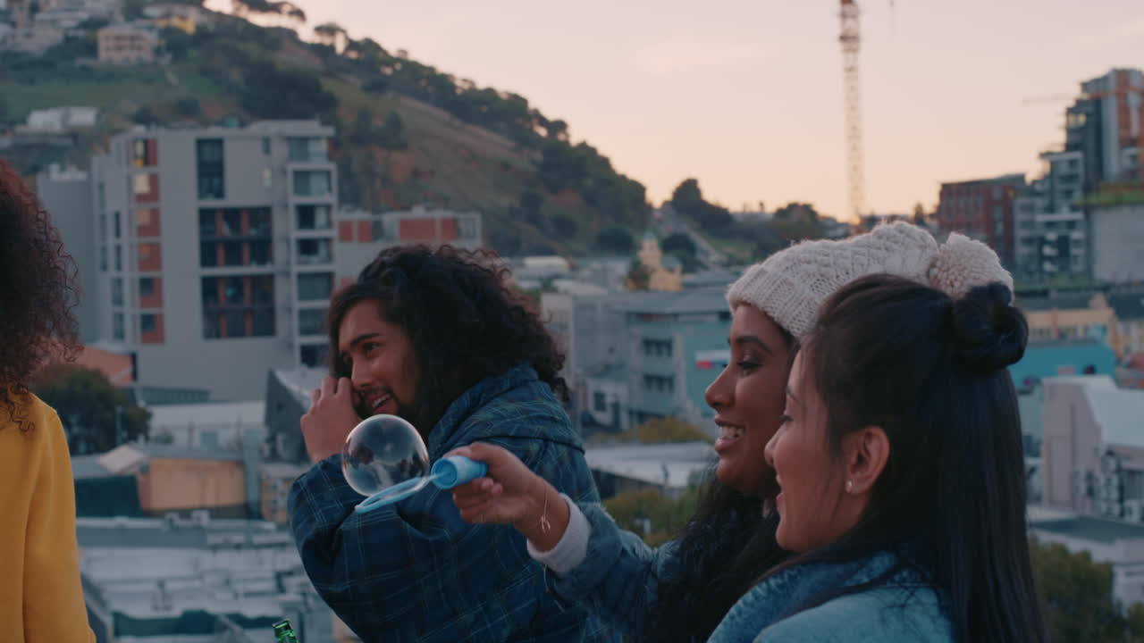 happy group of friends blowing bubbles on rooftop at sunset hanging out enjoying carefree weekend with urban city skyline in background