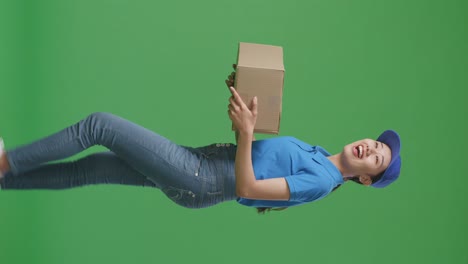 side view of smiling asian female courier in blue uniform walking and looking around while delivering a carton on green screen background in the studio