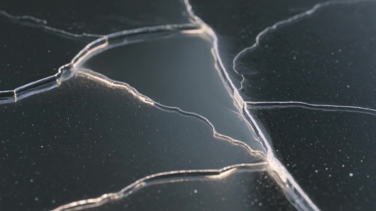 Triangular shard showing fracture tip advancing as low light grazes edges, glints on dark surface