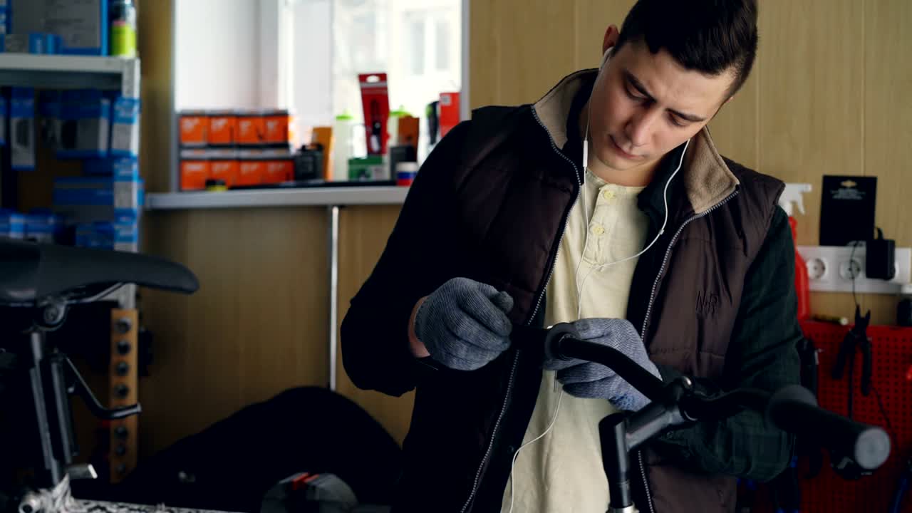Concentrated mechanic in gloves and warn vest is listening to music with earphones and fixing broken bicycle turning allen key in handle-bar. People and repair concept.