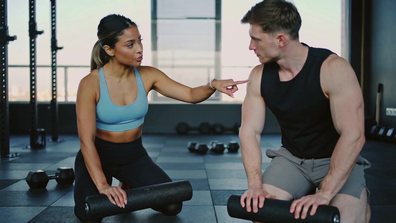 Zeigende Trainerin in blauem BH weist Mann im Fitnessstudio an, während dieser auf einer Faszienrolle seinen Rücken mobilisiert.