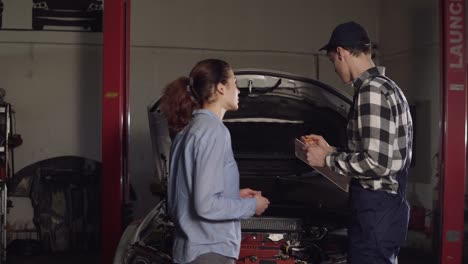 Niedlicher-Auto--Oder-Kfz-Techniker,-Der-Arbeitsmantel-Und-Mütze-Trägt-Und-Beschwerden-Seiner-Jungen-Kundin-In-Sein-Notizbuch-Schreibt