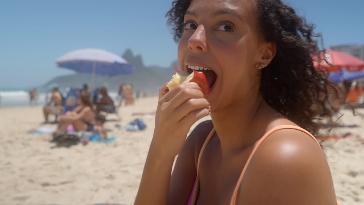 Nahaufnahme einer glücklichen multikulturellen Frau, die im Sand sitzt, einen roten Apfel isst und zu einem Rhythmus tanzt. Sie genießt den gesunden Snack unter der strahlenden Sonne am Strand von Ipanema
