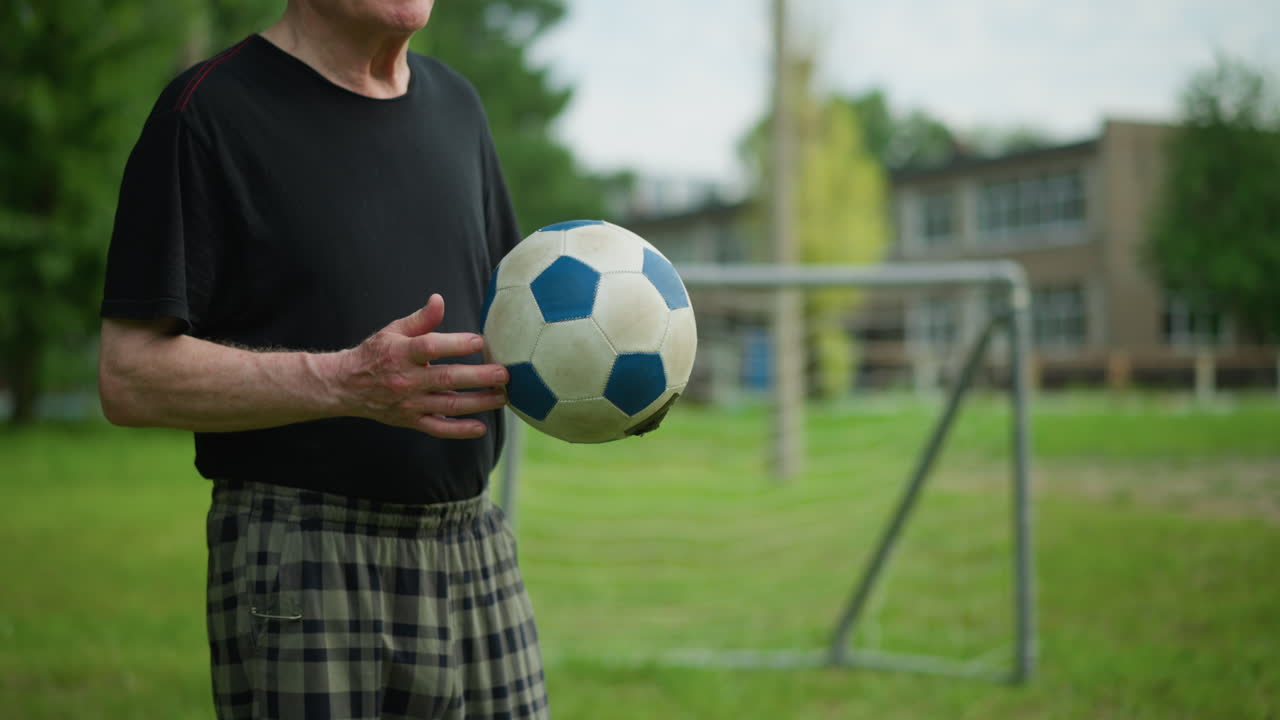 An elderly man rolls a soccer ball back and forth in his hand with focused intent, in the blurred background, a goalpost and a building can be seen
