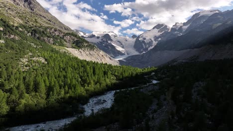 drone pushes in on lush green trees and stream with snowy mountain and cloudy sky in background at morteratsch glacier, val morteratsch, switzerland